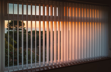 White vertical slat blinds hanging in front of a window as the sun is setting turning the light golden. The slats have sealed glued pockets and no cords at the bottom. - 307239926