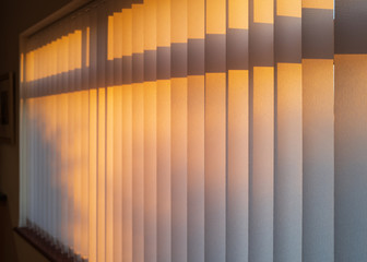 White textured vertical slat blinds hanging in front of a window as the sun is setting turning the...