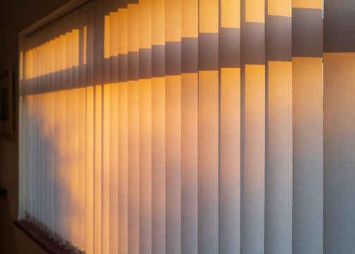 White Textured Vertical Slat Blinds Hanging In Front Of A Window As The Sun Is Setting Turning The Light Golden.