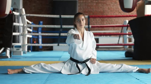 Lockdown of female martial arts fighter wearing white kimono with black belt sitting on floor in leg-split and doing punches