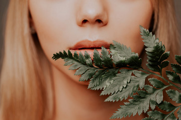 Close-up mouth of young woman with fresh healthy skin and hairs holding fern and smiling....