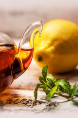 Glass teapot with tea on whitish boards with lemon and a sprig of fresh mint