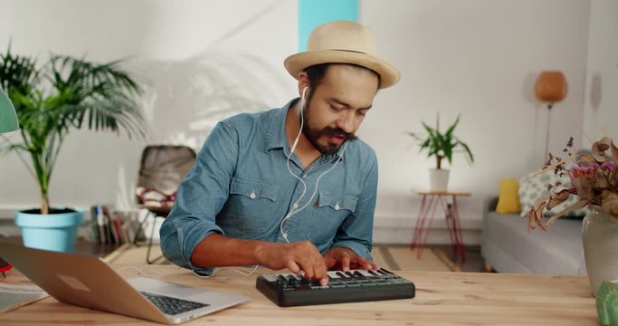 Cheerful Hispanic man creating music using silver laptop and midi controller at home desk, stylish guy in hat with beard and mustache dressed in hat and jeans shirt with white earphones in living room