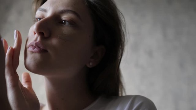 Close-up - caucasian woman rubs foundation smears on her cheekbone and looks in a small hand mirror. Anti age cream, lifting, smoothing face skin problems