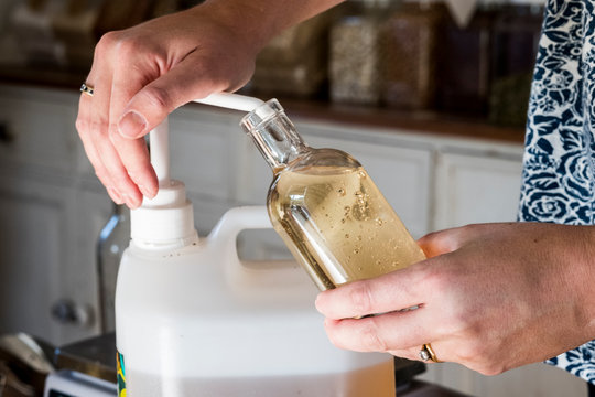 Close up of person standing in a kitchen, decanting liquid from plastic container into glass bottle.