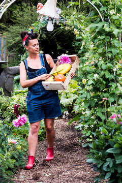 Woman walking in a garden, carrying wooden crate with fresh vegetables and flowers.