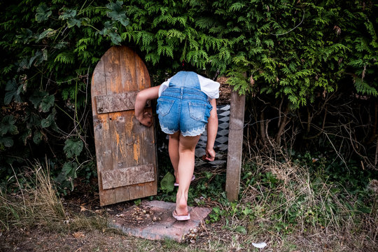 Rear view of girl wearing denim dungarees walking through small wooden door in a garden hedge.
