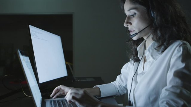 Teleworker talking and typing on laptop. Side view of young female call center operator in headset using computer at night. Customer service concept