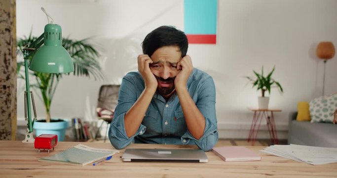 Tired melancholic young man feeling wailful, closing working laptop sitting behind table, lonely Hispanic guy with beard and mustache leaning at desk in cozy light living room, sad mood, bad thoughts