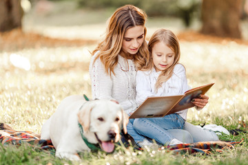 Fototapeta premium Happy beautiful mother and daughter with dog labrador is reads book sitting in the park.