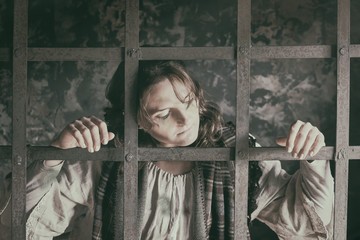 Girl in old clothes sad on the background of a prison cell and retro iron bars
