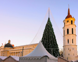 Fototapeta premium Night Cityscape with Christmas Market at Cathedral square in Winter Vilnius, Lithuania. Advent Fair Decoration and Stalls with Crafts Items on the Bazaar. Lithuanian street Xmas and holiday