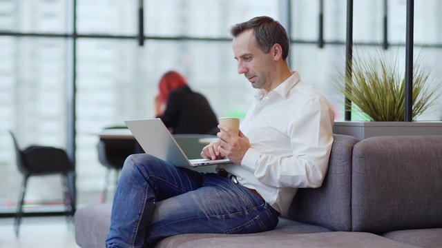 Medium tracking shot of middle aged businessman sitting on sofa in office lobby or coffee shop, drinking coffee and working on laptop computer, side view