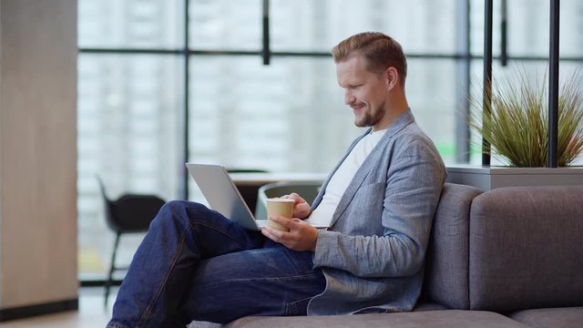 Tilt up medium shot side view of smiling middle aged man sitting on sofa in office lobby or coffee shop with coffee cup in his hand and browsing Internet on laptop computer