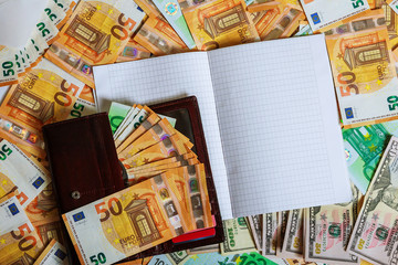 A white notebook with blank pages lay on a set of banknotes of 100 euros, 50 euros and 100 dollars.