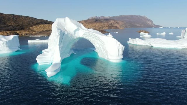 Slow and majestic drone aerial view over white and turquoise Icebergs floating in the calm Baffin Sea, Greenland, with rocky lands sitting in the background of this peaceful arctic landscape