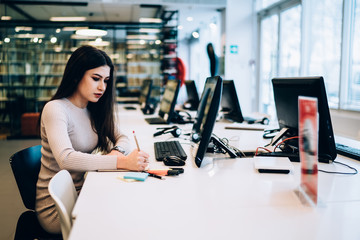 Focused female student using computer and writing in notebook while studying in library