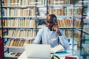 Serious ethnic student talking on smartphone near laptop in library