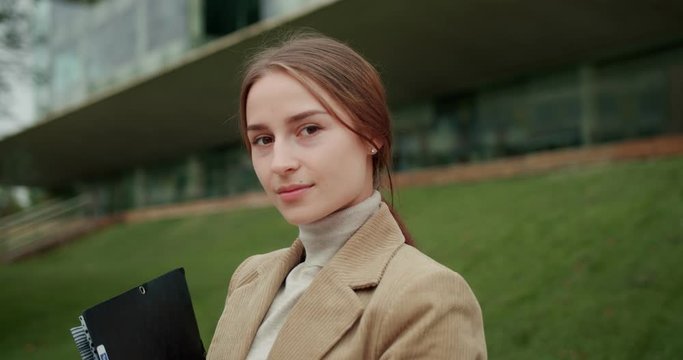 Portrait of self-confident successful Caucasian female turning face looking to camera, serious business woman holding folder with paper document outside modern office building dressed in jacket