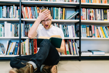 Shocked man reading book and touching forehead on floor near bookshelf