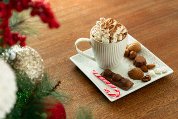 A cup of hot drink with marshmallows, candy stick, nuts, chocolate on wooden table, blurred Christmas tree