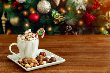 A cup of hot drink with marshmallows, candy stick, nuts, chocolate and caramel on wooden table, Christmas tree background with ornaments and lights, copy space