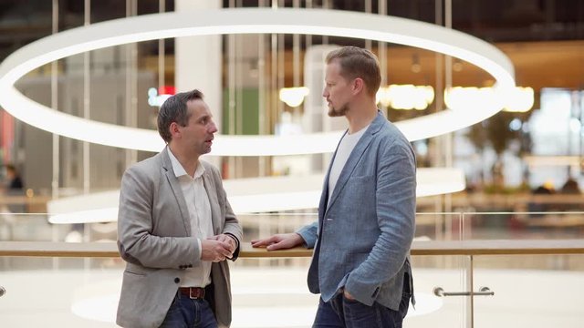 Medium tracking side view shot of two middle aged businessmen talking and discussing business issues while standing in office lobby leaning on railing