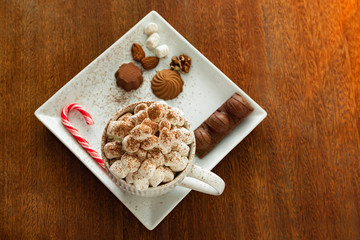 A cup of hot drink with marshmallows, candy cane, nuts, chocolate on wooden table, top view