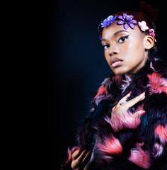 young pretty african american woman in spotted fur coat and flowers on head smiling on black background, fashion people concept