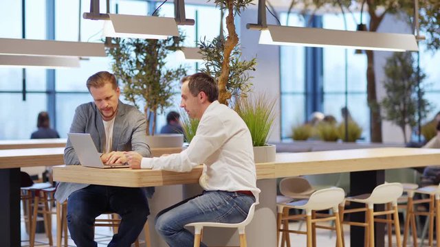 Tracking shot of two middle aged businessmen sitting at counter in cafeteria, watching and discussing business presentation on laptop screen