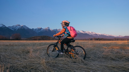 One caucasian children rides bike in wheat field. Little girl riding black orange cycle on background of beautiful snowy mountains. Biker motion ride with backpack and helmet. Mountain bike hardtail.