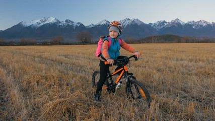 One caucasian children walk with bike in wheat field. Little girl walking black orange cycle on background of beautiful snowy mountains. Biker stand with backpack and helmet. Mountain bike hardtail.