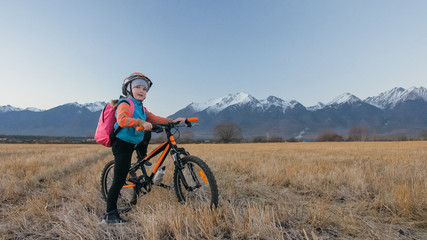 Obraz premium One caucasian children walk with bike in wheat field. Little girl walking black orange cycle on background of beautiful snowy mountains. Biker stand with backpack and helmet. Mountain bike hardtail.