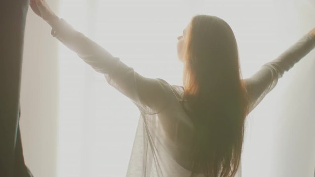 Beautiful Young Brunette Opens Curtains To let Morning Sunlight In. Following Back View Of Attractive Girl Rising Early and Greeting New Day Happily