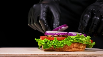 Craft burger is cooking on black background in black food gloves. Consist: red sauce salsa, lettuce, red onion, pickle, cheese, chilli green pepper, air bun and marbled meat beef. Not made ideal