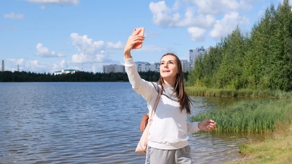 Beautiful girl with a backpack takes a selfie near the lake and talks on a cell phone on Skype.