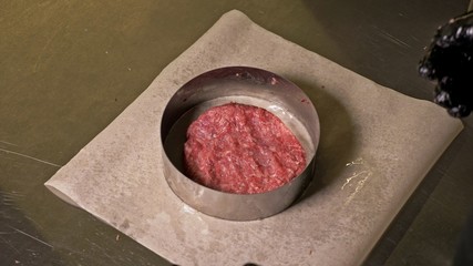 Cook makes meat medallions for burgers. Chef in black food gloves makes cutlet. Cutlets are leveled in steel ring in an even medallion. On top of pouring spices pepper and salt.