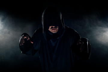 Man dressed in a dark robe looking like a cult leader on a smoky or foggy background.  He looks...