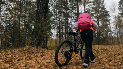 Fototapeta premium One caucasian children walk with bike in autumn park. Little girl walking black orange cycle in forest. Kid goes do bicycle sports. Biker motion ride with backpack and helmet. Mountain bike hardtail.