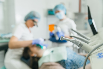 Naklejka na ściany i meble Woman at dentist clinic gets dental treatment to fill a cavity in a tooth. Dental restoration and composite material polymerization with UV light and laser. Doctors in the background are out of focus.