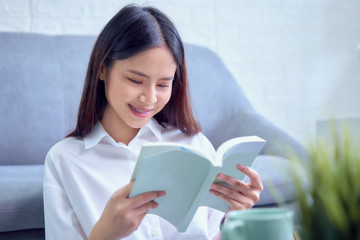 Fototapeta premium Happy young Asian woman sitting on sofa and reading book in her living room, vacation time.