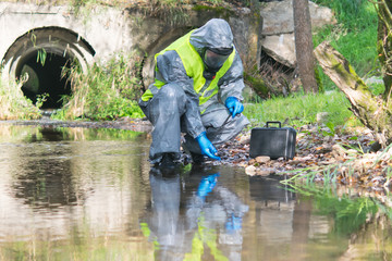 a specialist in a protective suit and mask, is measuring the device on the pond, holding it in his hands, against the background of the treatment pipe