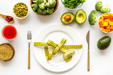 Diet concept. Fork and knife tied with measure tape on plate near vegetables on white background top view