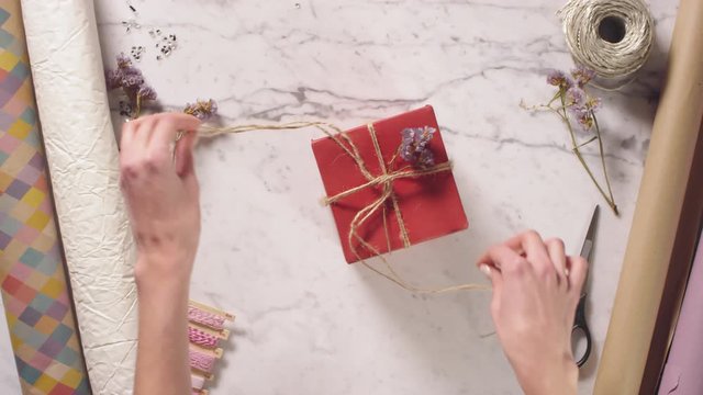 Top view shot of hands of woman removing jute rope and decorative dried flower from red gift box, opening it and taking bottle of perfume while unpacking present on marble table