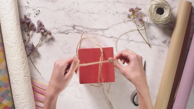 Top view fast motion shot of female hands packing bottle of perfume into red gift box, wrapping jute rope around, tying a bow and decorating present with dried flower on white marble table