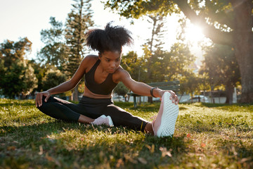 Portrait of an african american female athlete stretching her legs sitting on lawn in the park - young black woman warming up her muscles before exercising 