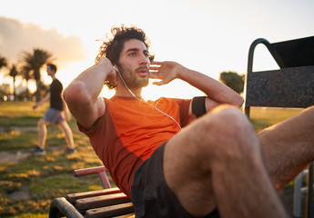 Confident young man listening to music in headphones from smart phone armband doing exercises on public equipment in the outdoor gym at the park - obrazy, fototapety, plakaty