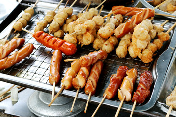 Fried sausages and meatballs in bamboo skewers on aluminum tray with selective focus. A roadside restaurant is a cart.