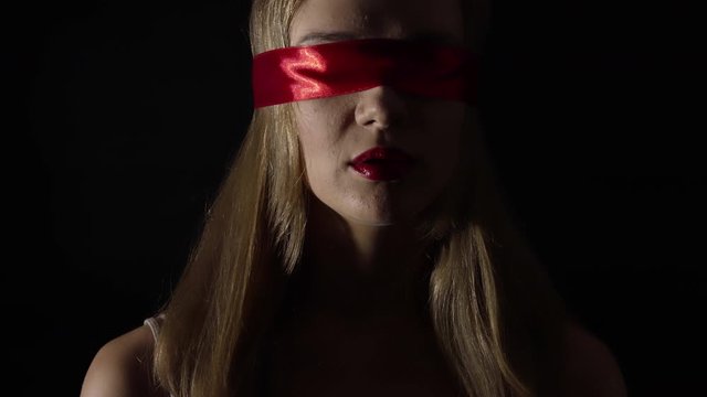 Portrait of the young woman blindfold with red ribbon over black background. Fetish concept.
