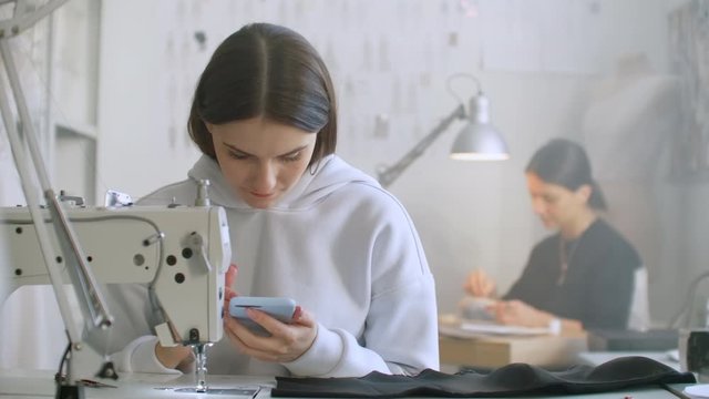 Woman seamstress looks at the smartphone screen in the workplace. a colleague works and sews on a sewing machine. A break in the work of the seamstresses. Internet banking during work